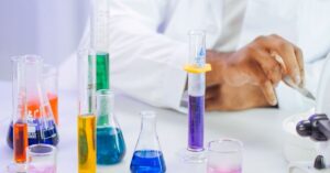 Tangan seorang ilmuwan bekerja dengan cairan berwarna di dalam suasana laboratorium.